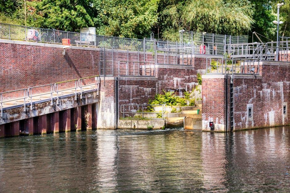 A riverside scene in a garden area along Brentford Lock shows a brick retaining wall with metal railings and steps leading down to the water. The brickwork varies in color, with some sections appearin